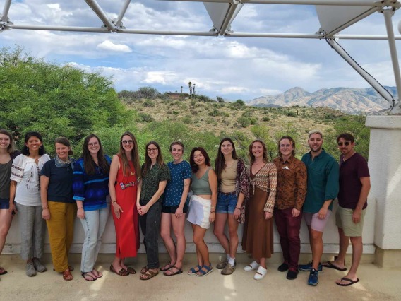 Photo of Carson Scholars 2023-2024 cohort. 13 graduate students standing in a line at Biosphere 2 with the Sonoran Desert in the background.