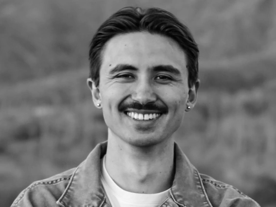Black and white image of Tai Koester: Man with short black hair and a mustache wearing a white shirt and jacket. His arms are crossed, and he is smiling in front of a mountain.