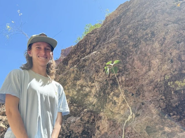 Molly Gans - woman with shoulder length hair wearing a white shirt and grey pants. She is standing next to a wall of earth with some plants growing on it. 