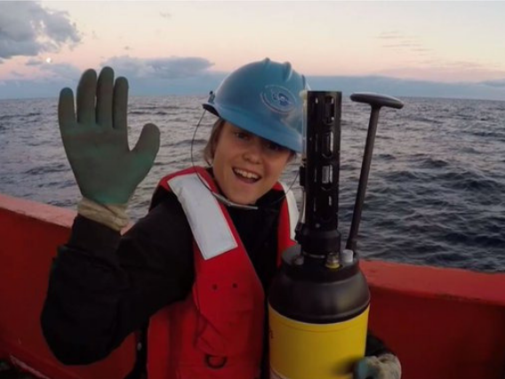 Image of Becki Beadling holding a large sensor with a blue hardhat on standing on a large red boat in the arctic ocean 