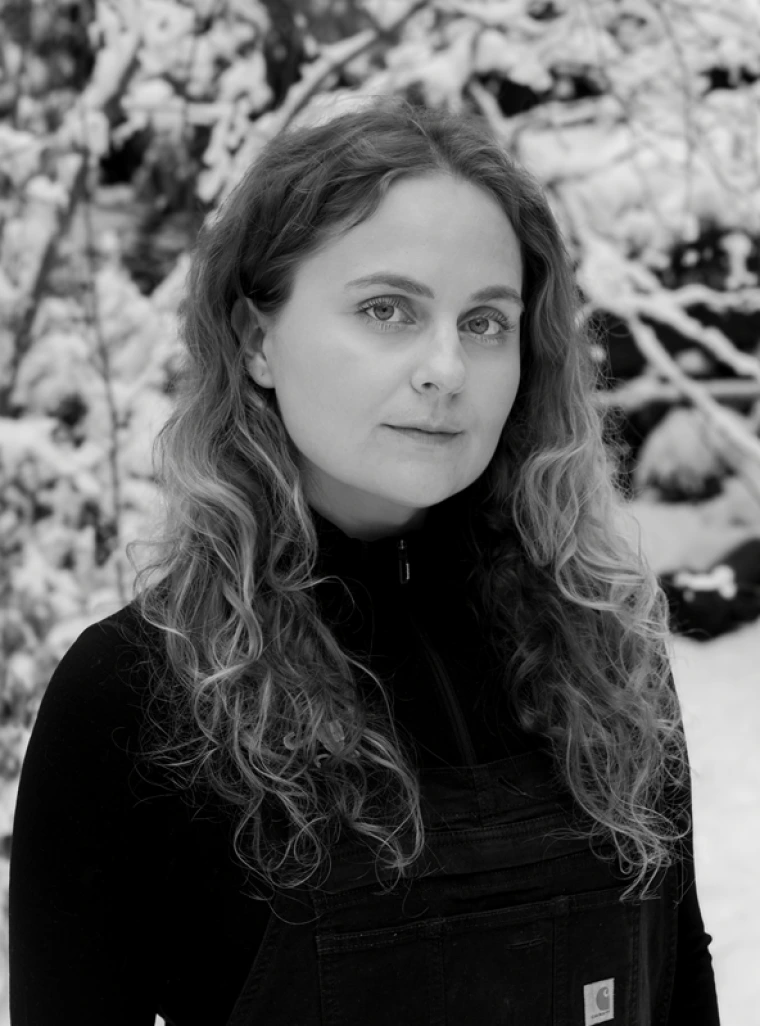 Black and white photo of woman with long, wavy hair looking at the camera. She is wearing a black sweater and standing in front of snowy branches.