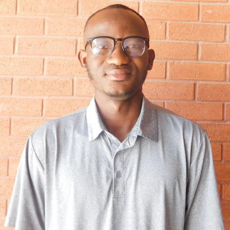 Prosper Babon-Ayeng - a man with very short hair, light facial hair, and square glasses standing against a brick wall. He is wearing a grey polo shirt. 