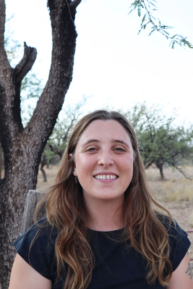 Madeleine Wallace - a woman with long brown hair wearing a blue shirt smiling at the camera and standing in front of a tree