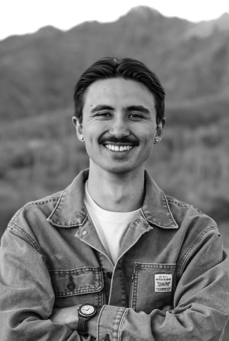 Black and white image of Tai Koester: Man with short black hair and a mustache wearing a white shirt and jacket. His arms are crossed, and he is smiling in front of a mountain.