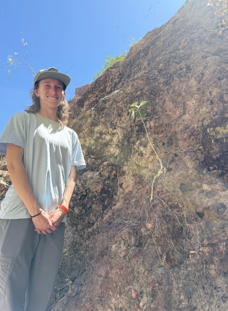 Molly Gans - woman with shoulder length hair wearing a white shirt and grey pants. She is standing next to a wall of earth with some plants growing on it.