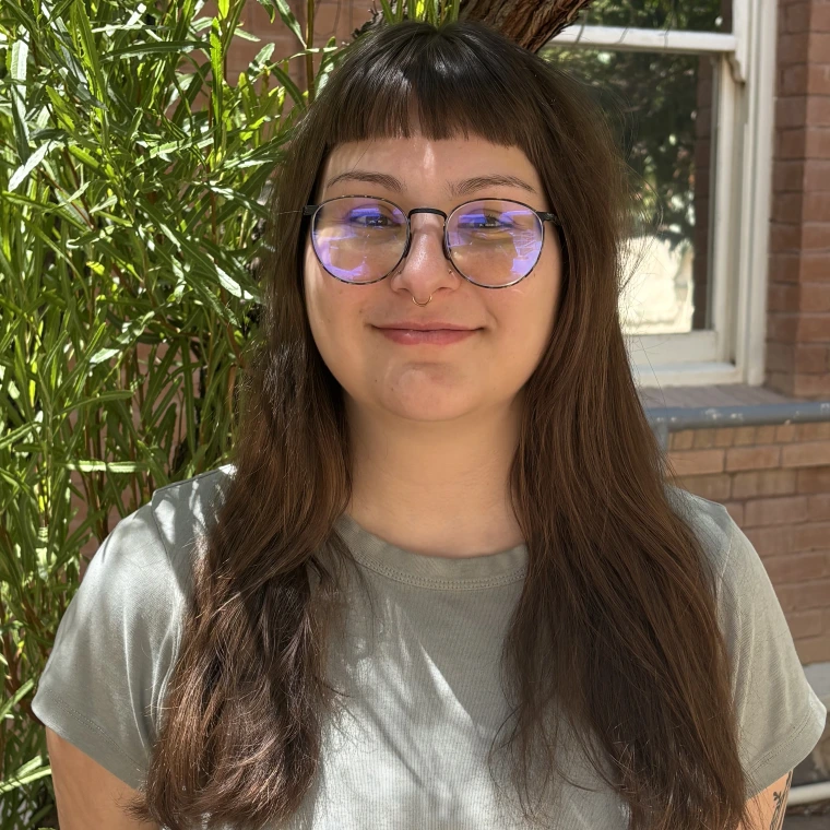A person with long brown hair and short bangs is wearing a grey shirt and round glasses while smiling at the camera. They are standing in front of a brick building with some greenery behind them.