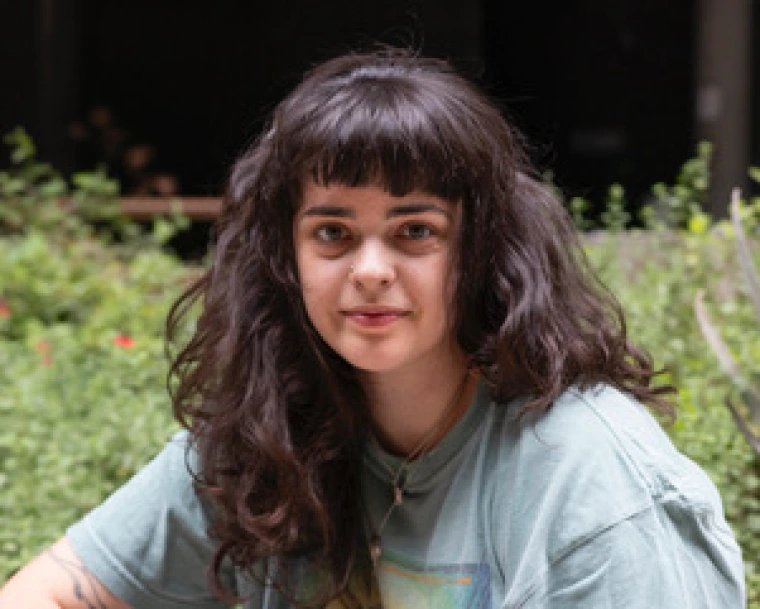 A woman with thick, wavy, brown hair and blunt bangs smiles at the camera. She is sitting in front of greenery and wearing a grey shirt. 