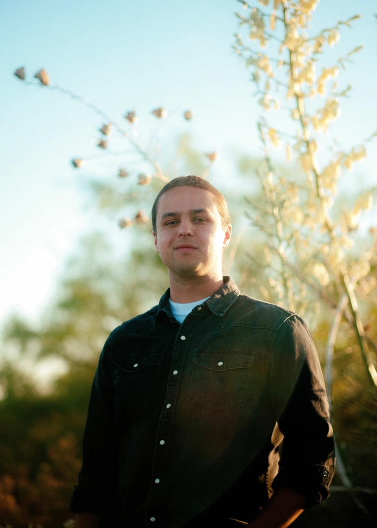 A man with pulled back brown hair stands in front of a tall plant and blue sky. He is wearing a dark green button-up shirt.