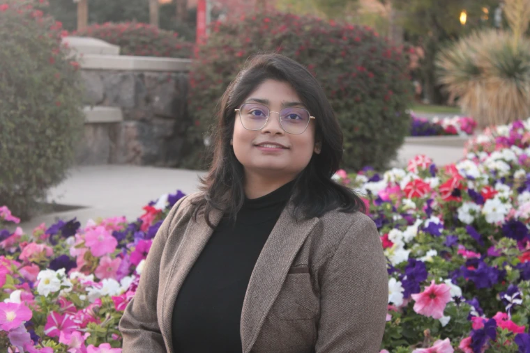 A woman with chest-length black hair and round wire glasses smiles at the camera. She is standing in front of a garden with flowers and bushes. She is wearing a black shirt and beige blaiser. 