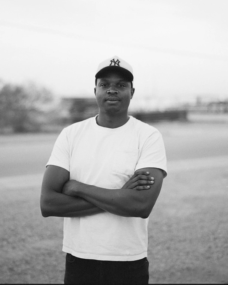 Black and white photo of a man standing in front of a blurred background. His arms are crossed, and he is wearing a white t-shirt and baseball cap.