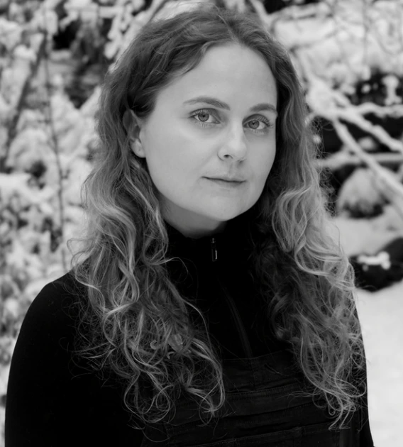 Black and white photo of woman with long, wavy hair looking at the camera. She is wearing a black sweater and standing in front of snowy branches.